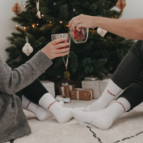Zwei Personen stoßen mit Glühwein an, tragen „Team Glühwein“-Socken, Weihnachtsbaum im Hintergrund.