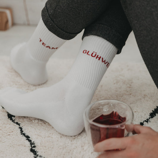 Person in weißen „Team Glühwein“-Socken sitzt mit einem Glas Glühwein auf einem hellen Teppich.