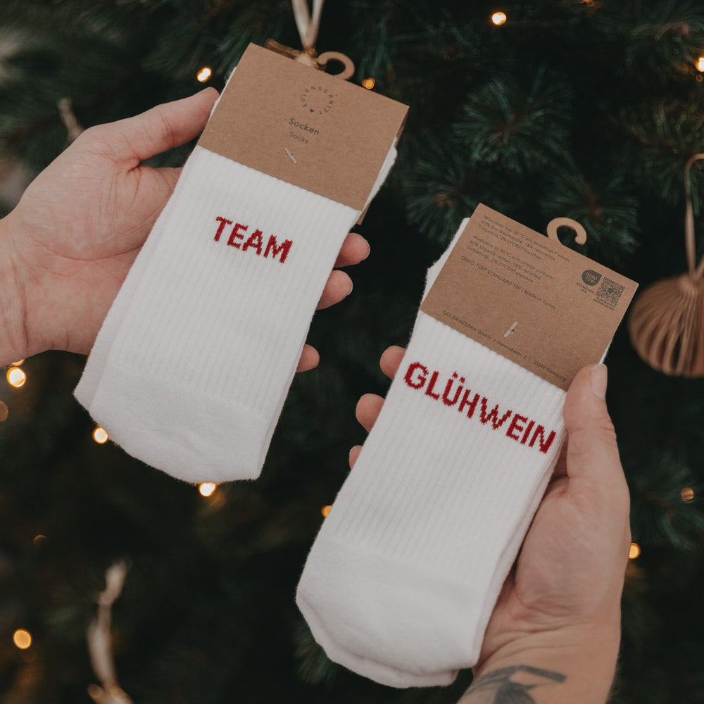Zwei Hände halten Socken mit rotem Schriftzug „Team Glühwein“ vor einem geschmückten Weihnachtsbaum.