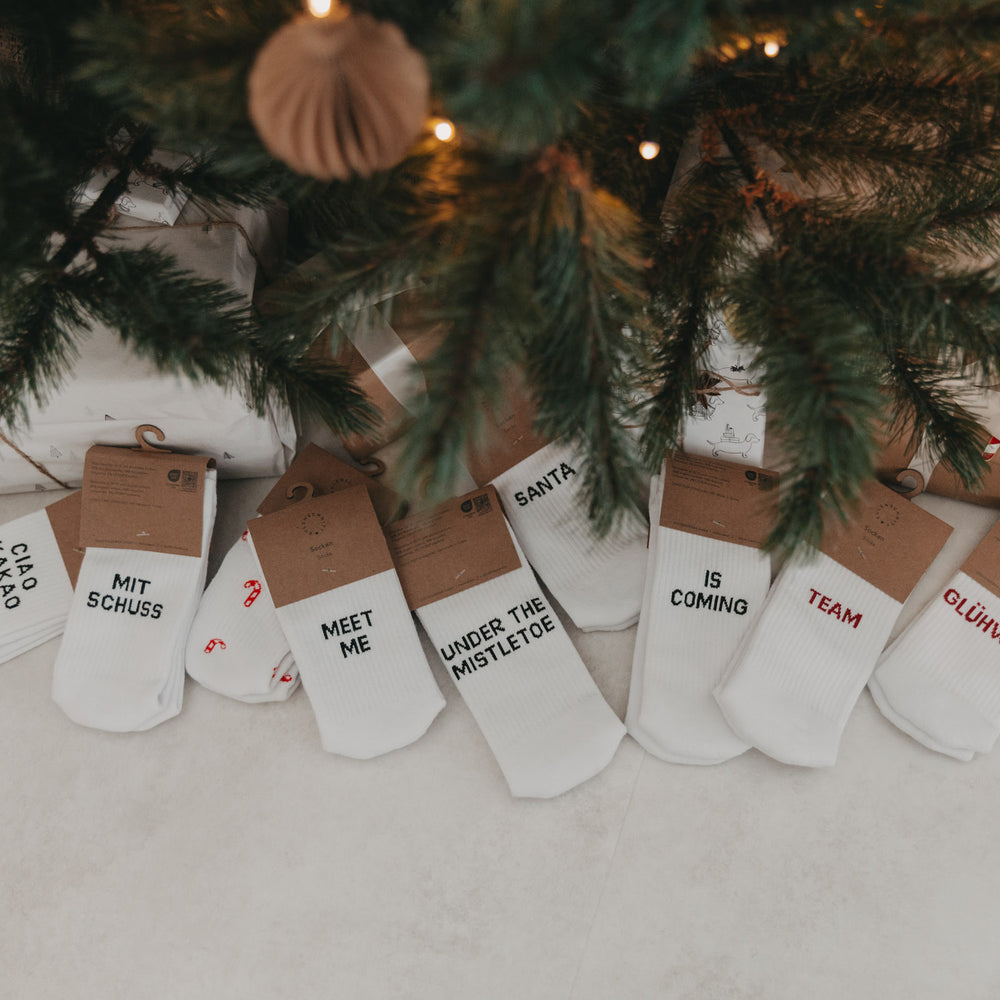 Mehrere Paar Eulenschnitt-Socken unter einem Weihnachtsbaum – darunter „Team Glühwein“ und „Santa is coming“.