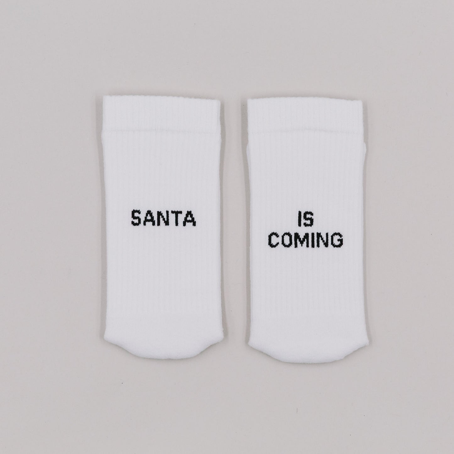Weiße Socken mit schwarzem Schriftzug „Santa is coming“ nebeneinander auf hellem Hintergrund