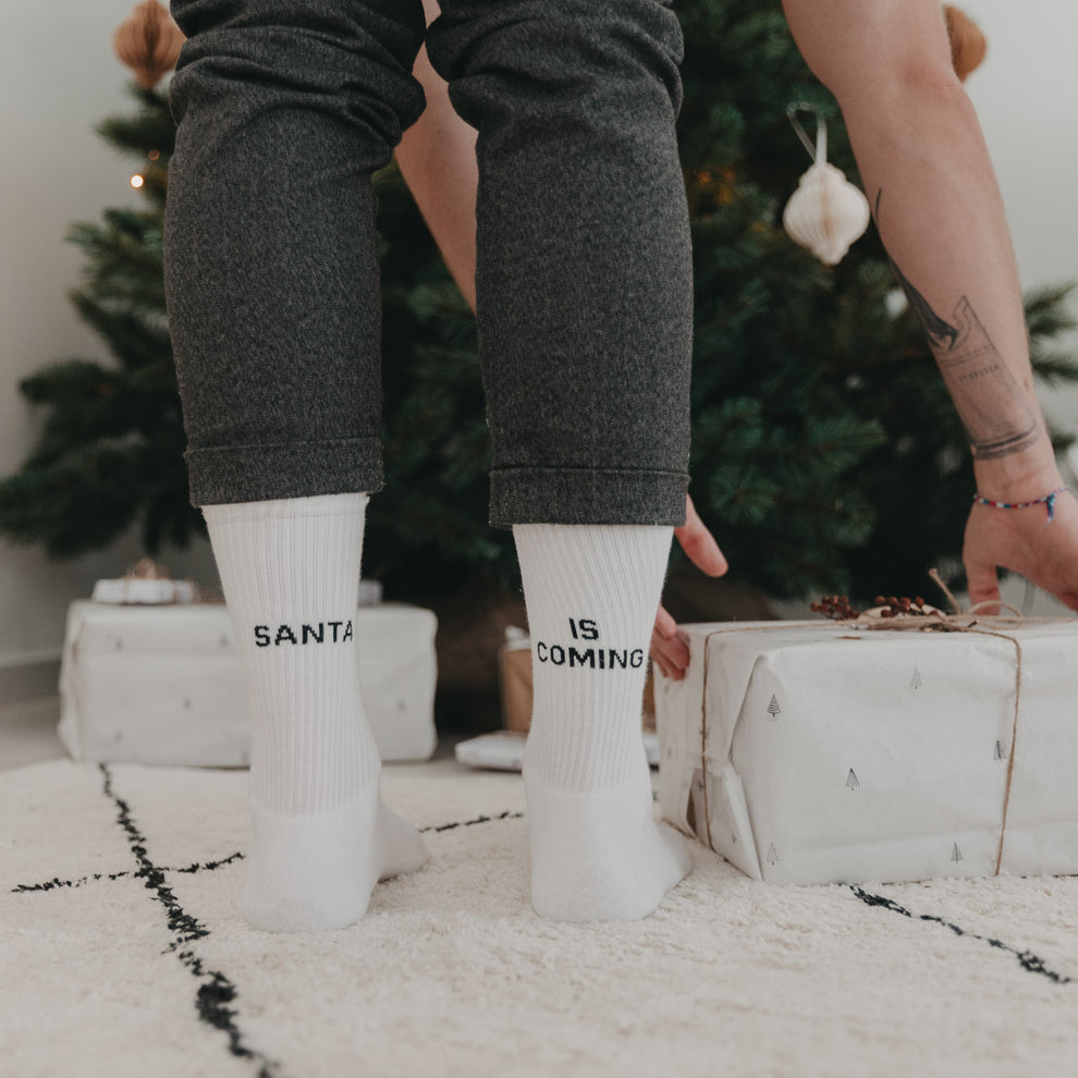 Person mit weißen „Santa is coming“-Socken beim Geschenkeplatzieren vor geschmücktem Tannenbaum