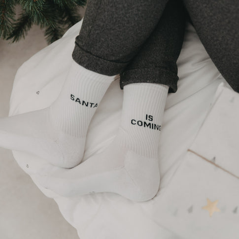 „Santa is coming“-Socken an gemütlich ausgestreckten Füßen auf Bettdecke mit Weihnachtsdeko