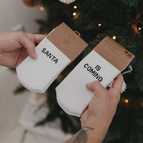 Nahaufnahme der verpackten „Santa is coming“-Socken mit Kartonverpackung vor Weihnachtsbaum