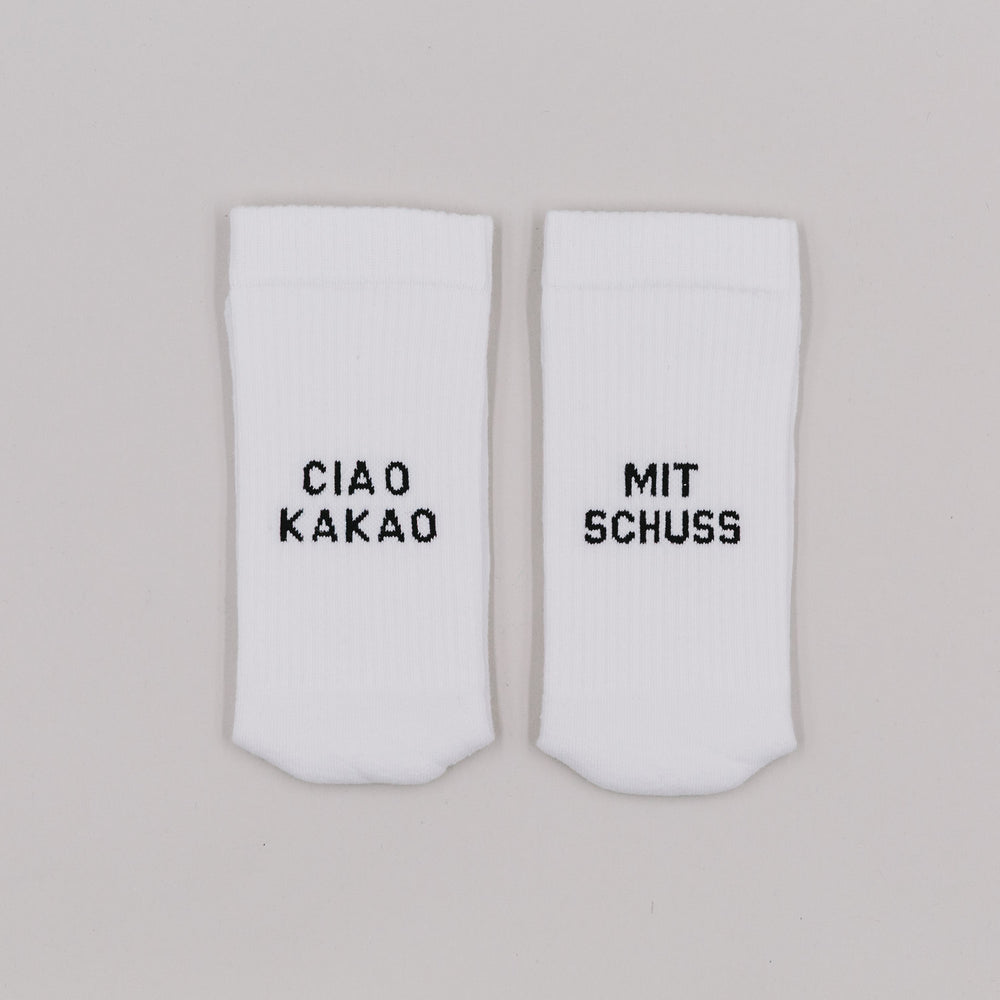 Weiße Socken mit schwarzer Stickerei „Ciao Kakao“ und „Mit Schuss“ auf neutralem Untergrund nebeneinander liegend