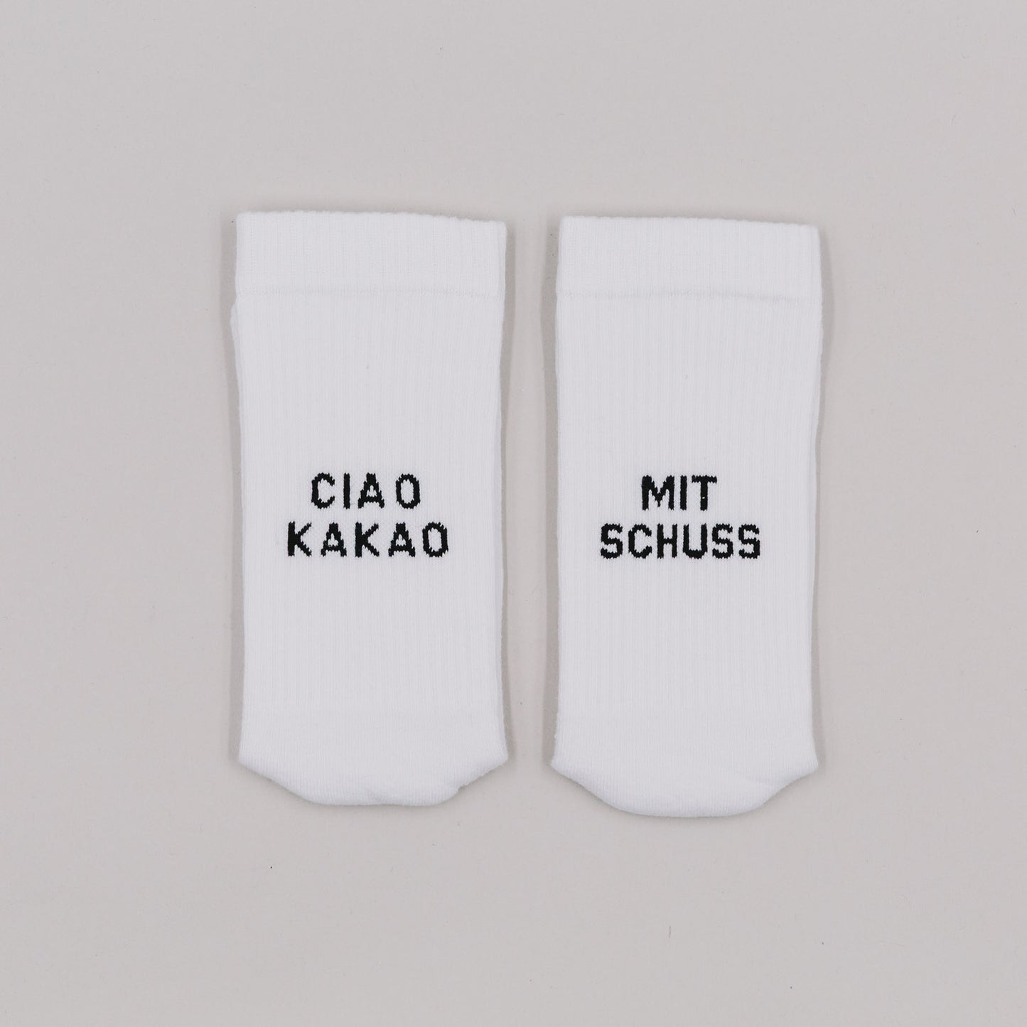 Weiße Socken mit schwarzer Stickerei „Ciao Kakao“ und „Mit Schuss“ auf neutralem Untergrund nebeneinander liegend