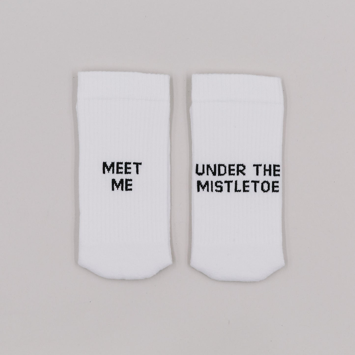 Weiße Socken mit der Aufschrift „Meet me“ und „Under the mistletoe“ auf grauem Hintergrund nebeneinander liegend