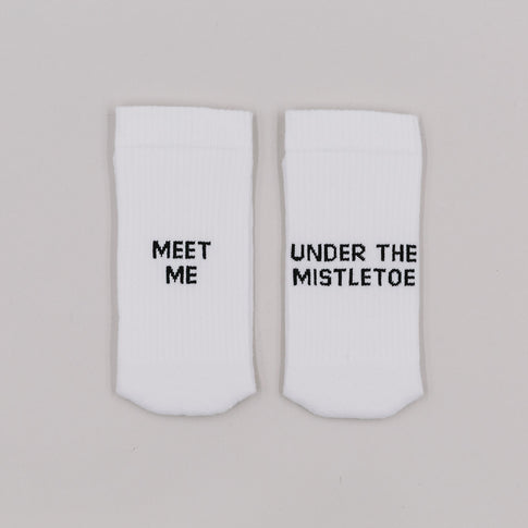 Weiße Socken mit der Aufschrift „Meet me“ und „Under the mistletoe“ auf grauem Hintergrund nebeneinander liegend