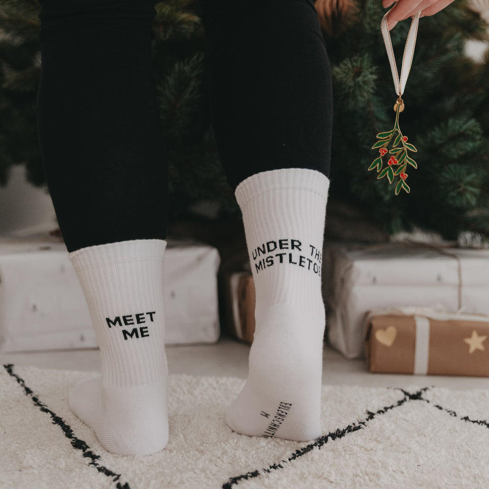 Person mit weißen Socken „Meet me under the mistletoe“ steht vor einem Weihnachtsbaum, in der Hand ein Mistelzweig