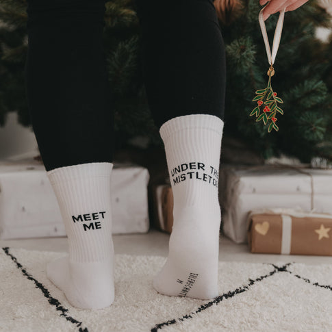 Person mit weißen Socken „Meet me under the mistletoe“ steht vor einem Weihnachtsbaum, in der Hand ein Mistelzweig