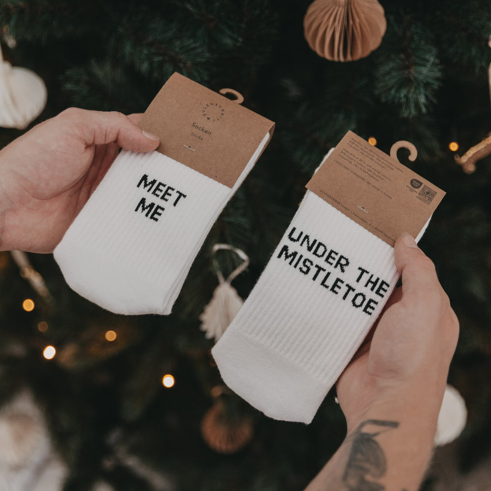 Socken „Meet me“ und „Under the mistletoe“ in Verpackung vor einem dekorierten Weihnachtsbaum in Händen gehalten