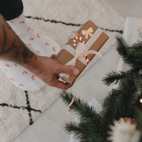 Zuckerstangen-Socken im Hintergrund, Hand hält Geschenk vor geschmücktem Weihnachtsbaum.