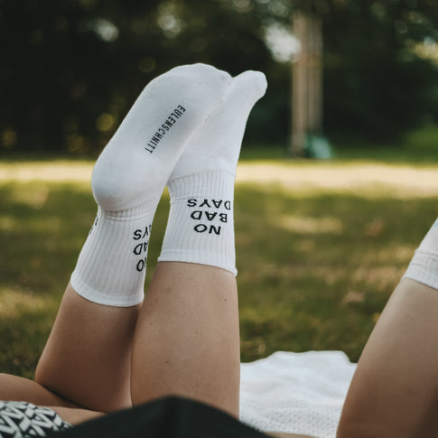 Nahaufnahme: Eine Person liegt auf einer Decke im Gras, ihre Füße mit weißen NO BAD DAYS-Socken zeigen in die Luft, die Markenaufschrift Eulenschnitt ist auf den Sohlen zu sehen.