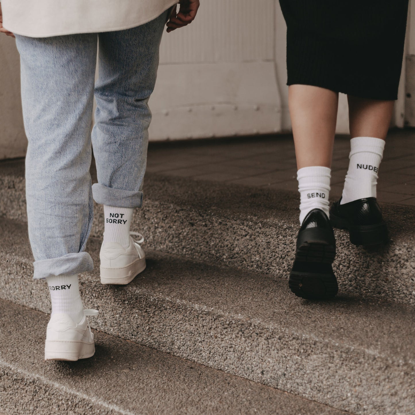 Zwei Mädels von hinten beim Gehen – beide tragen weiße Socken mit schwarzem Statement-Schriftzug, darunter „SEND NUDES“ und „SORRY NOT SORRY“.