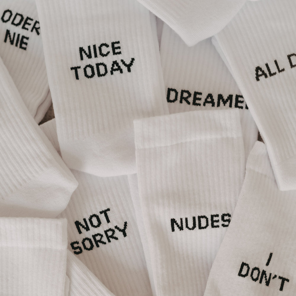 Mehrere weiße Statement-Socken mit schwarzer Aufschrift, gestapelt und überlappend – zu lesen sind u.a. „NICE TODAY“, „NOT SORRY“, „NUDES“, „I DON’T“ und „DREAMER“.