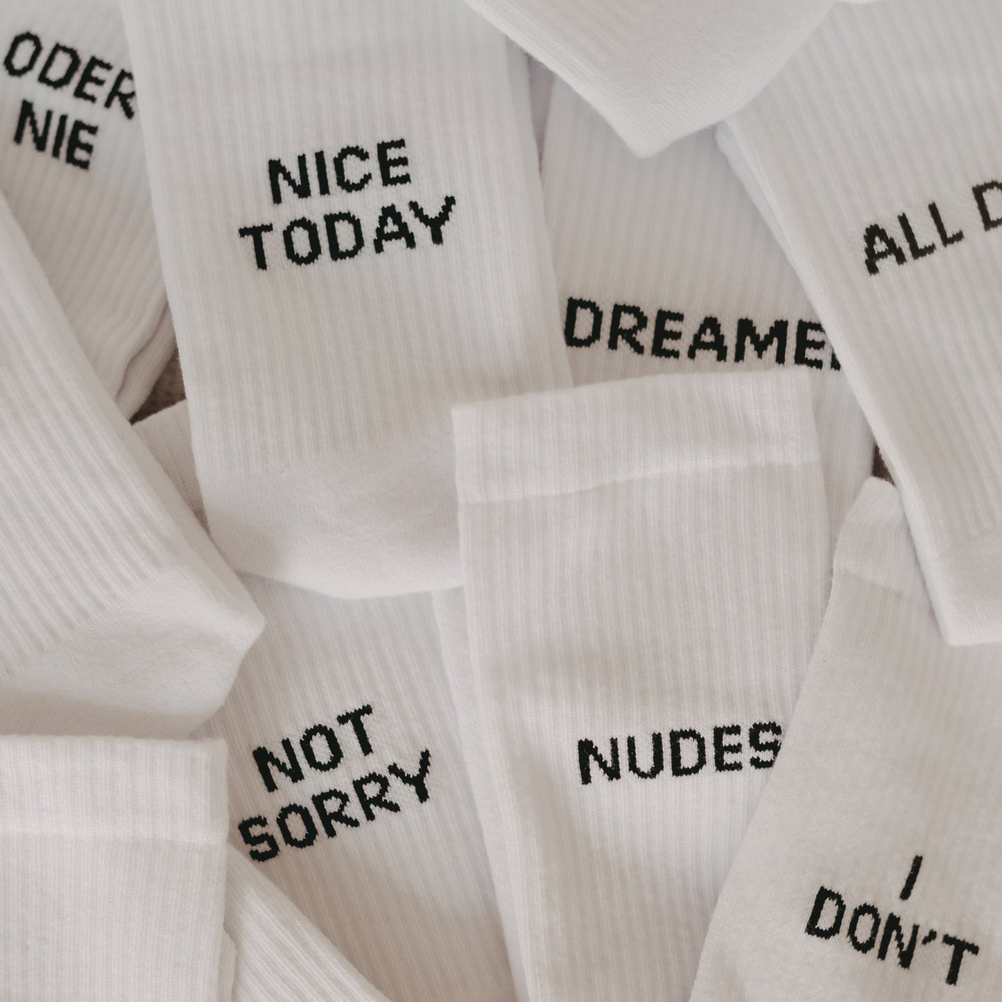 Mehrere weiße Statement-Socken mit schwarzer Aufschrift, gestapelt und überlappend – zu lesen sind u.a. „NICE TODAY“, „NOT SORRY“, „NUDES“, „I DON’T“ und „DREAMER“.