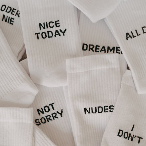 Mehrere weiße Statement-Socken mit schwarzer Aufschrift, gestapelt und überlappend – zu lesen sind u.a. „NICE TODAY“, „NOT SORRY“, „NUDES“, „I DON’T“ und „DREAMER“.