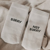 Weiße sportliche Socken mit schwarzem Schriftzug „SORRY“ und „NOT SORRY“ liegen auf einer beigen Decke.