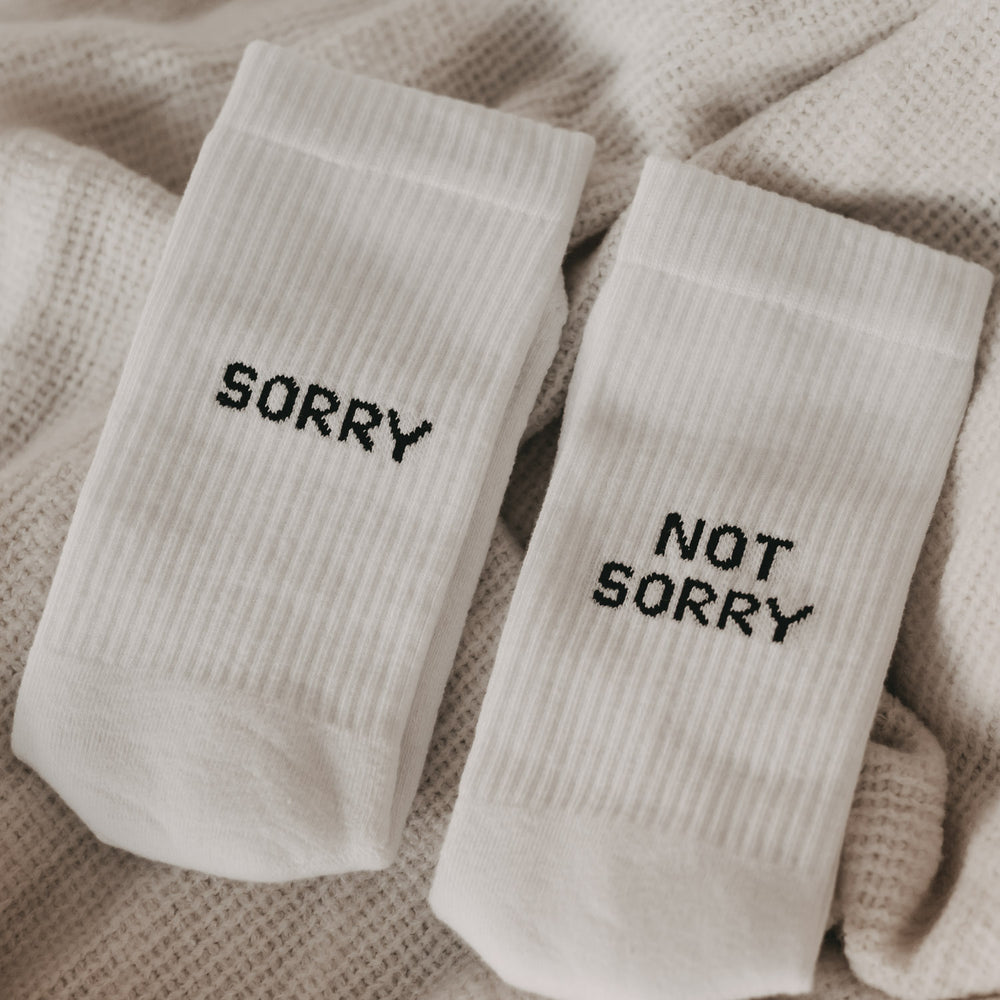 Weiße sportliche Socken mit schwarzem Schriftzug „SORRY“ und „NOT SORRY“ liegen auf einer beigen Decke.