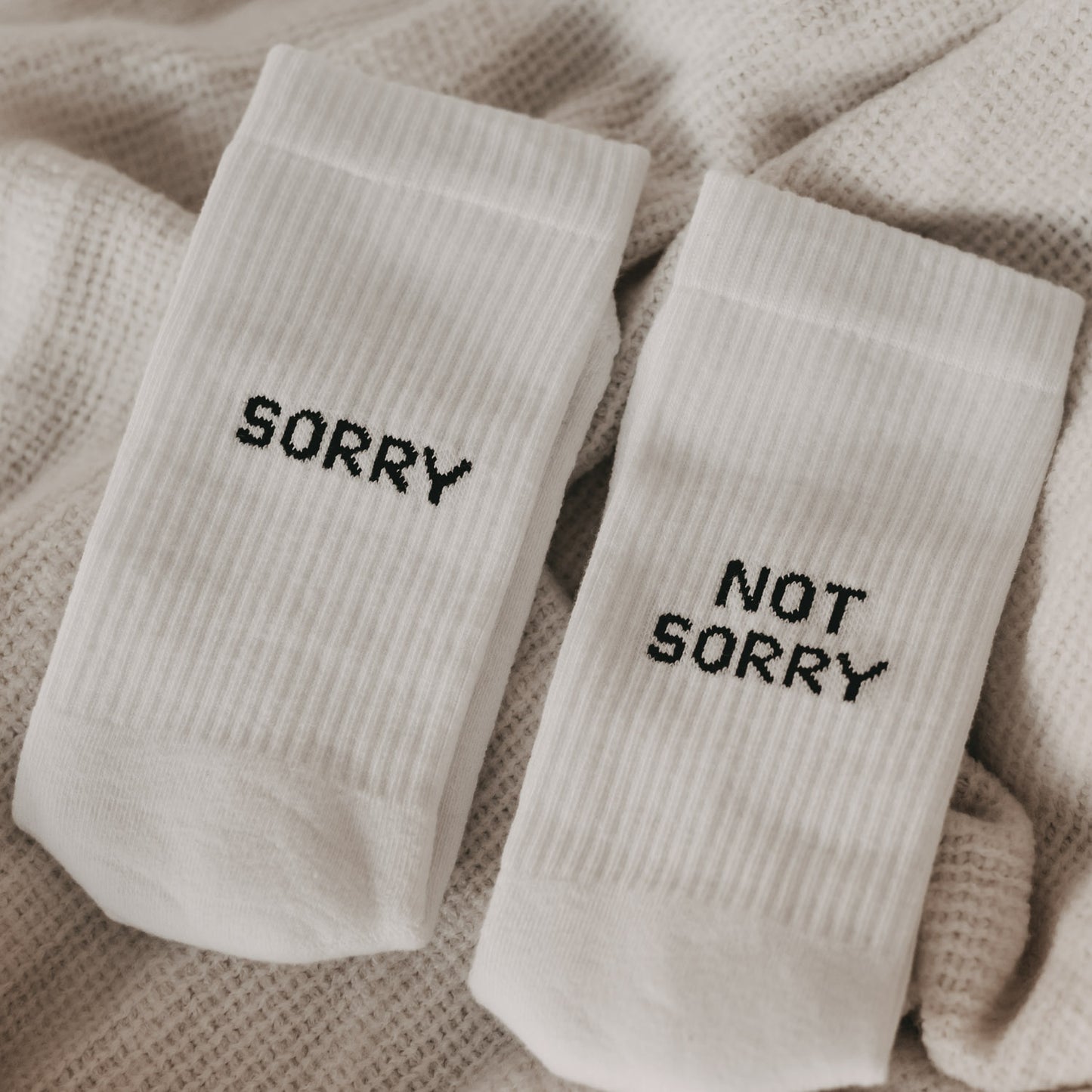 Weiße sportliche Socken mit schwarzem Schriftzug „SORRY“ und „NOT SORRY“ liegen auf einer beigen Decke.