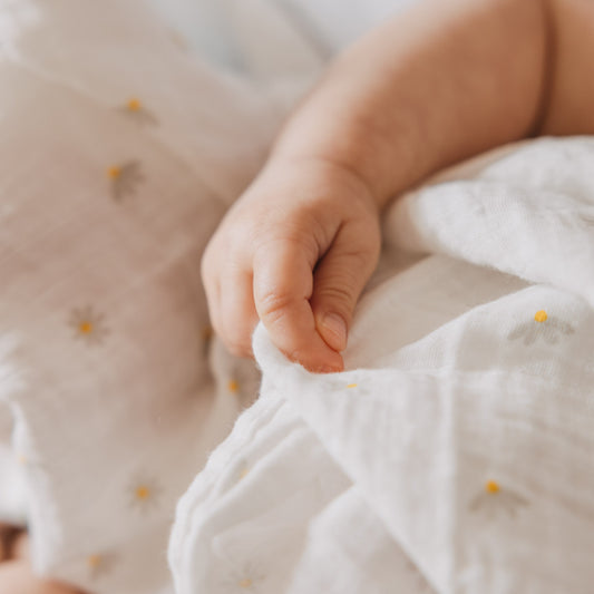 Kleine Babyhand greift nach einem weichen Musselintuch mit zartem Gänseblümchen-Print. Die Nahaufnahme hebt die feine Struktur und die angenehme Haptik des Stoffes hervor.