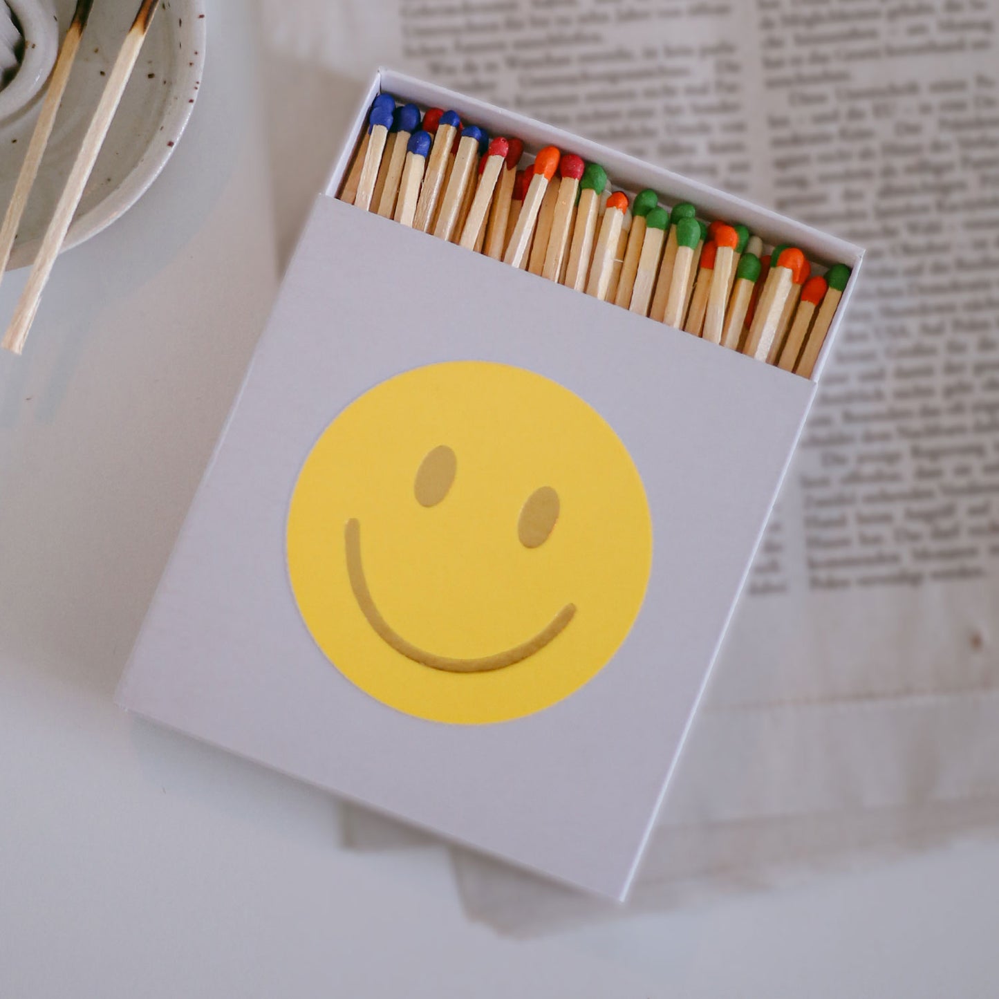 Streichholzschachtel mit gelbem Smiley und bunten Streichhölzern auf Tisch mit Zeitung.