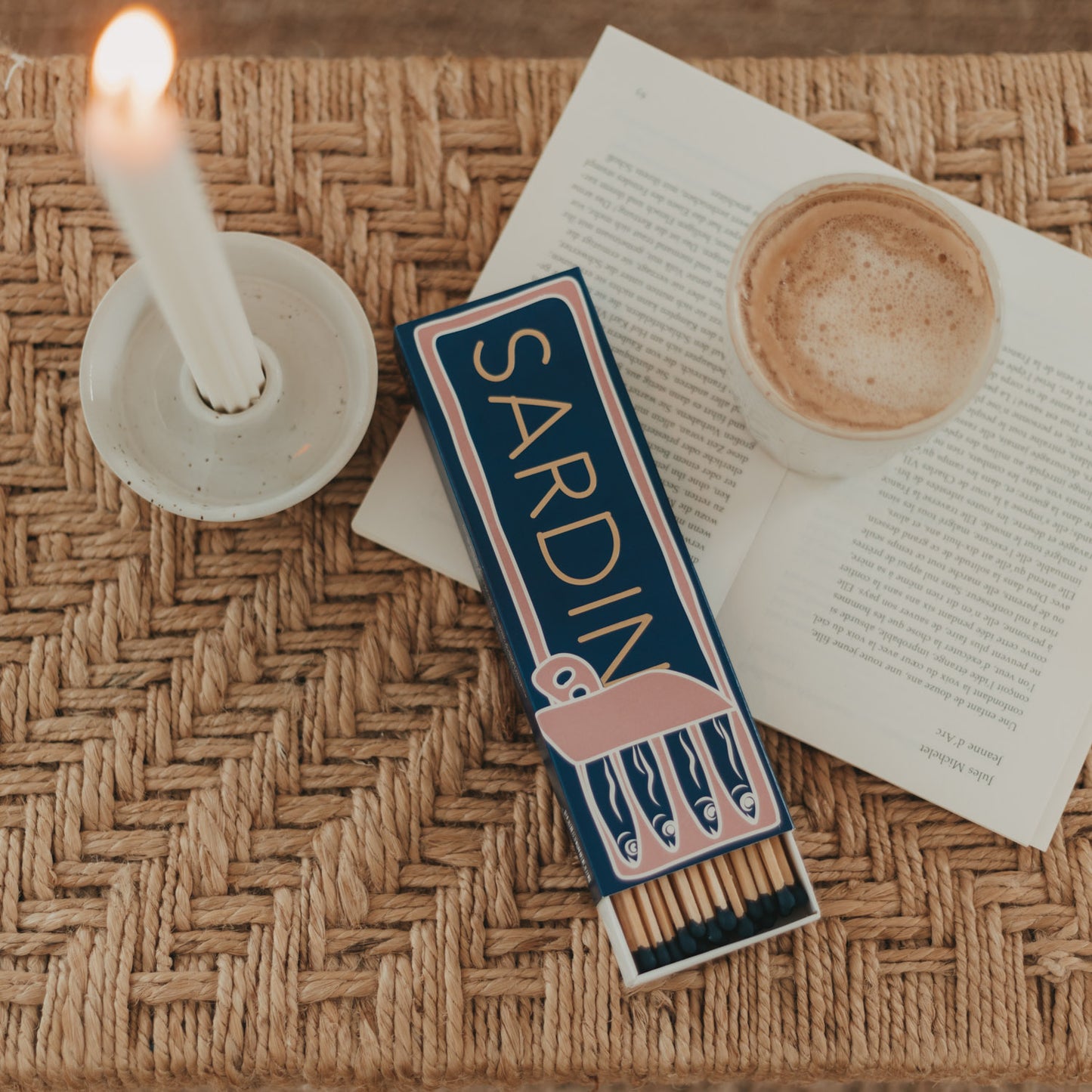 Streichholzschachtel „Sardinen“ in Blau und Rosa mit langen Streichhölzern neben Kerze und Kaffee