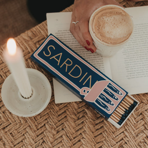 Geöffnete Streichholzschachtel „Sardinen“ mit blauen Streichholzköpfen neben Kerze und Kaffeetasse