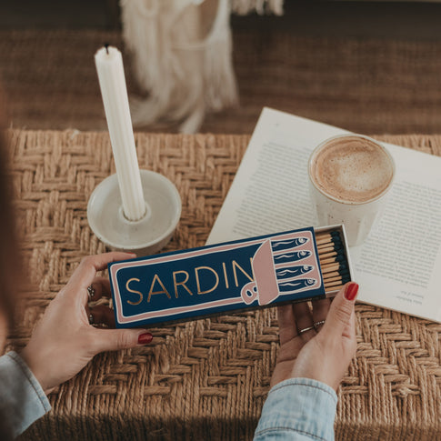 Streichholzschachtel „Sardinen“ mit langen Streichhölzern in den Händen neben Kerze und Kaffee