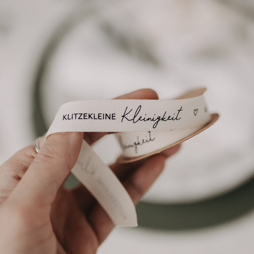 Hand hält weißes Stoff-Geschenkband mit Aufdruck „Klitzekleine Kleinigkeit“.