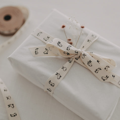 Weiße Geschenkbox mit Kaninchen-Geschenkband und Trockenblumen auf hellem Tisch.