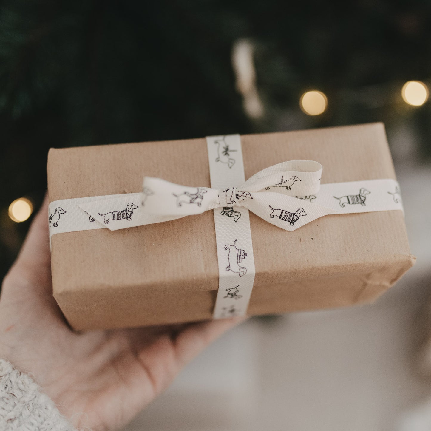 Hand hält Geschenk mit Baumwollband, Dackelmotiv mit Schal und Pullover, Weihnachtsbaum im Hintergrund