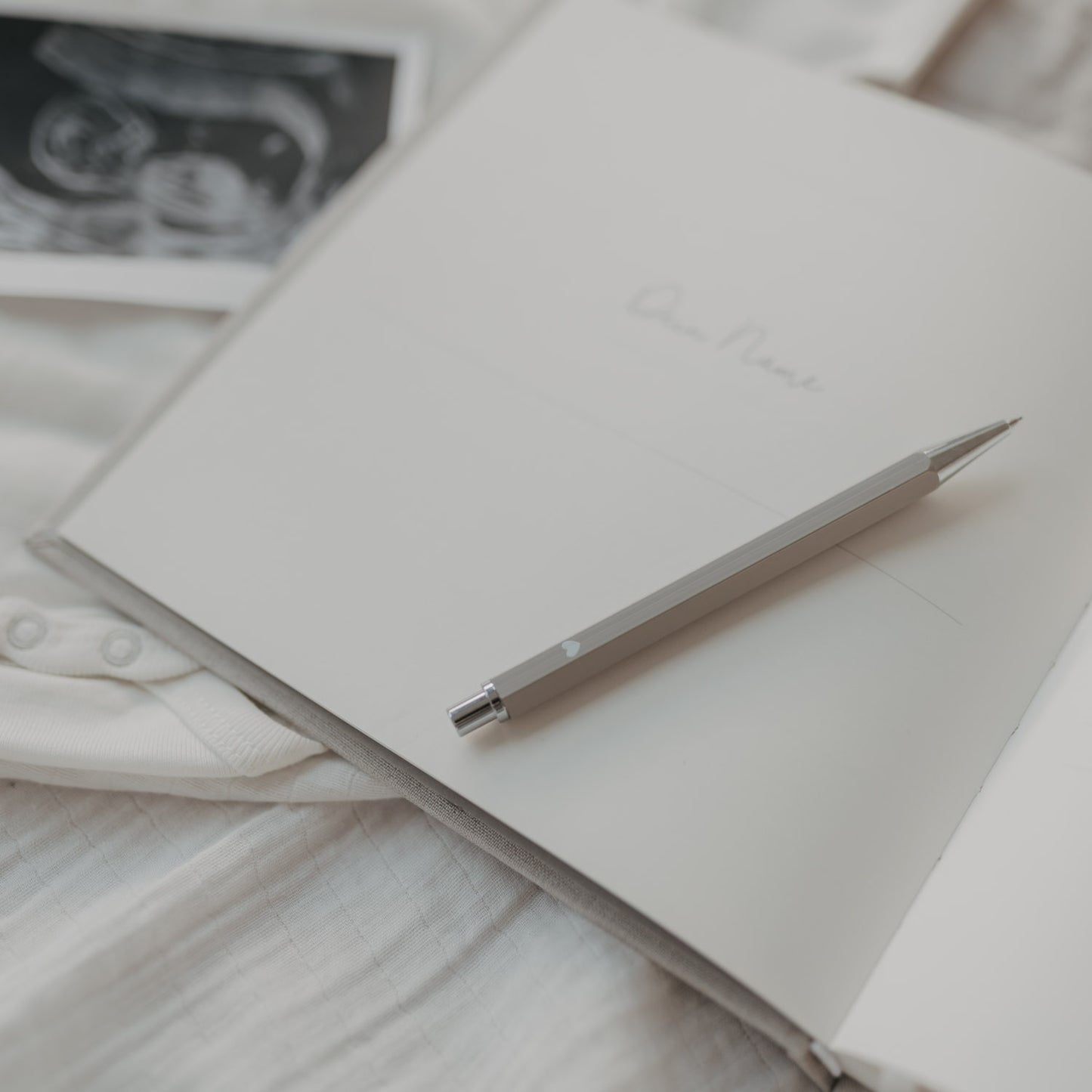 Grauer Druckbleistift auf offenem Notizbuch, umgeben von sanften Stoffen und Ultraschallbild.