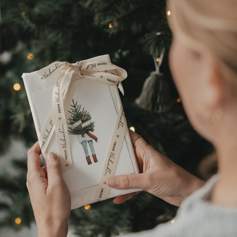 Weihnachtsgeschenk mit Eulenschnitt Geschenkband Frohe Weihnachten in den Händen vor dem Weihnachtsbaum