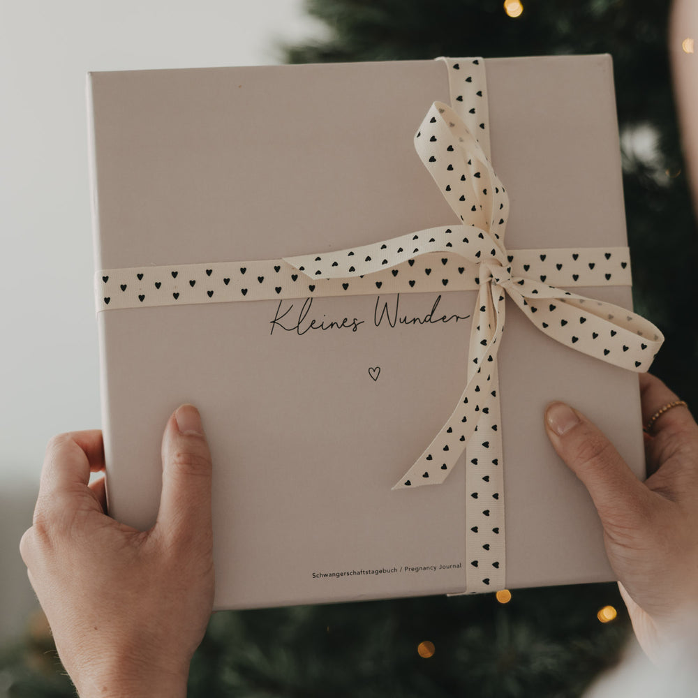 Geschenk mit beigem Geschenkband und schwarzen Herzen vor einem Weihnachtsbaum