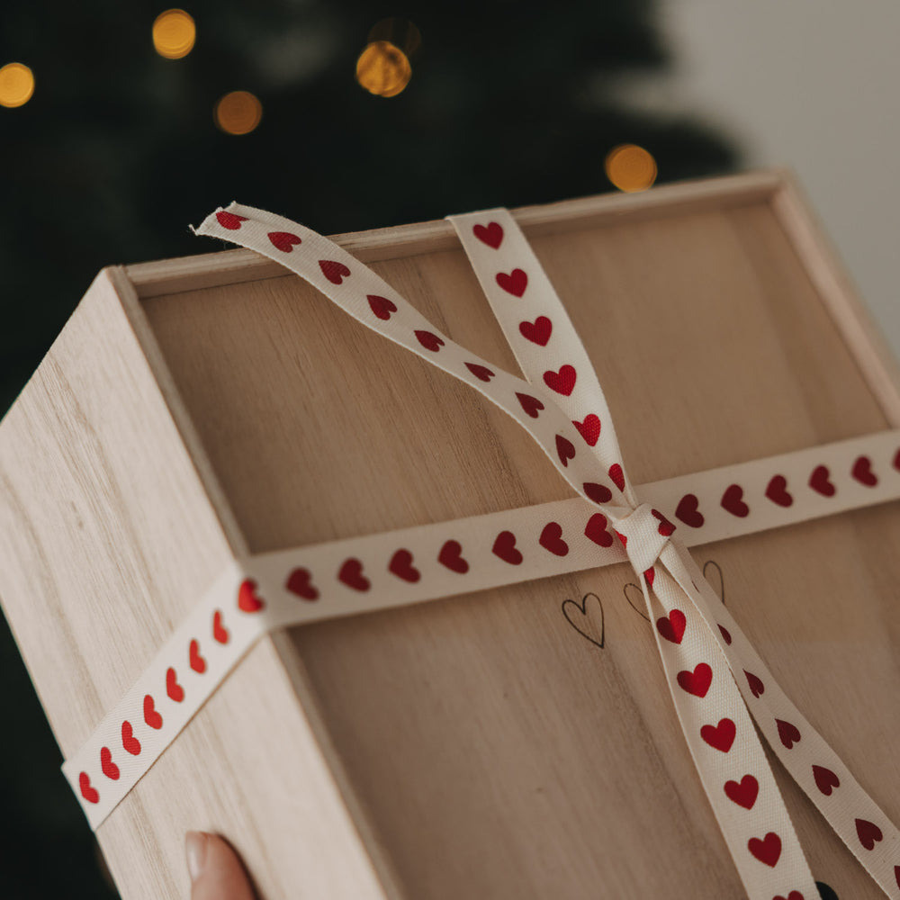 Holzbox mit beigem Geschenkband und roten Herzen vor einem Weihnachtsbaum