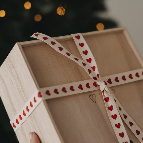 Holzbox mit beigem Geschenkband und roten Herzen vor einem Weihnachtsbaum