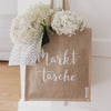 Jutetasche mit weißem Schriftzug „Markttasche“, gefüllt mit weißen Hortensien und einem gepunkteten Tuch, hängt an einer Tür.