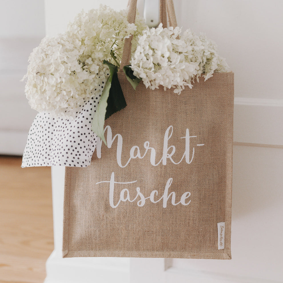 Jutetasche mit weißem Schriftzug „Markttasche“, gefüllt mit weißen Hortensien und einem gepunkteten Tuch, hängt an einer Tür.