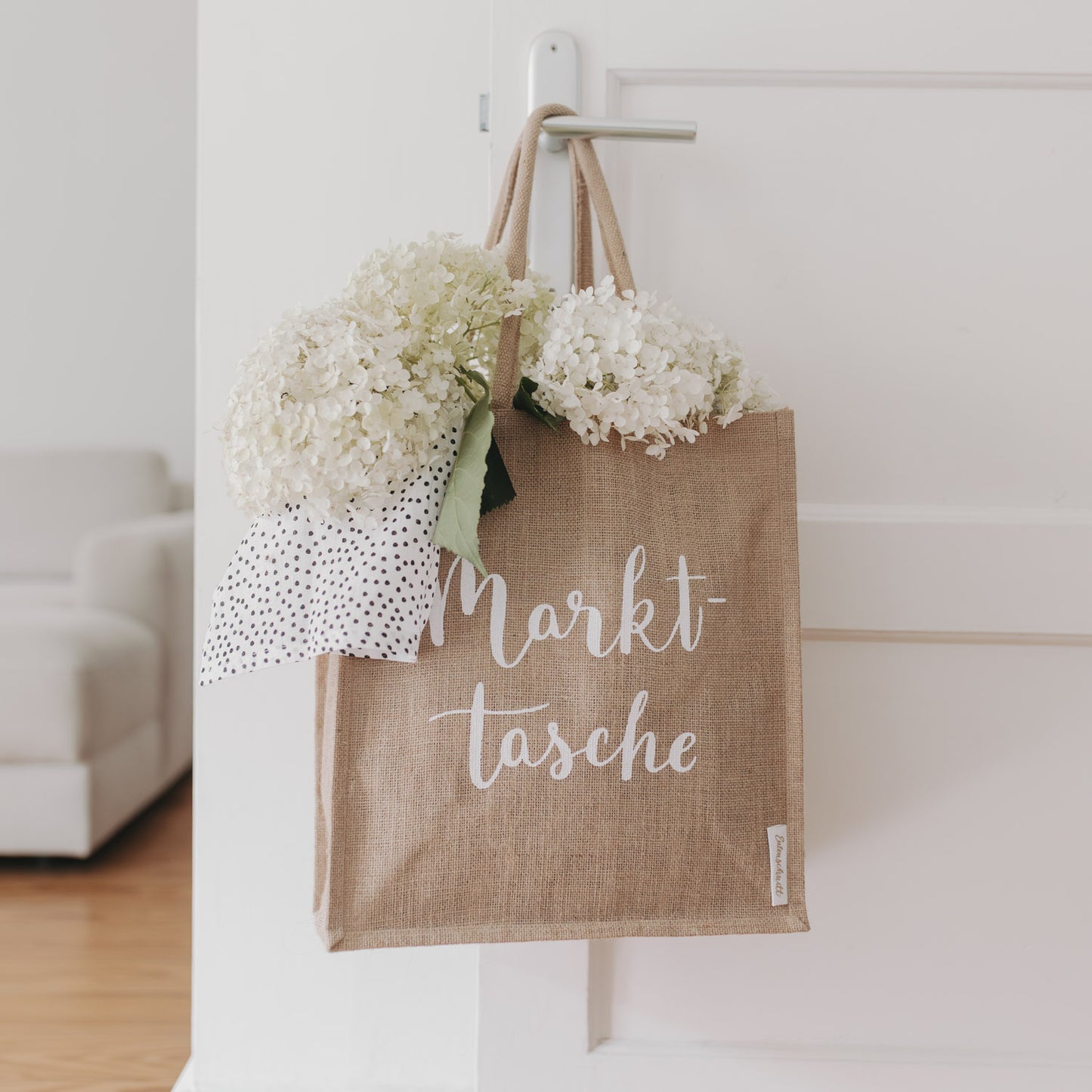 Die „Markttasche“ hängt allein an einem weißen Haken, befüllt mit weißen Blüten – schlicht und elegant in Szene gesetzt.