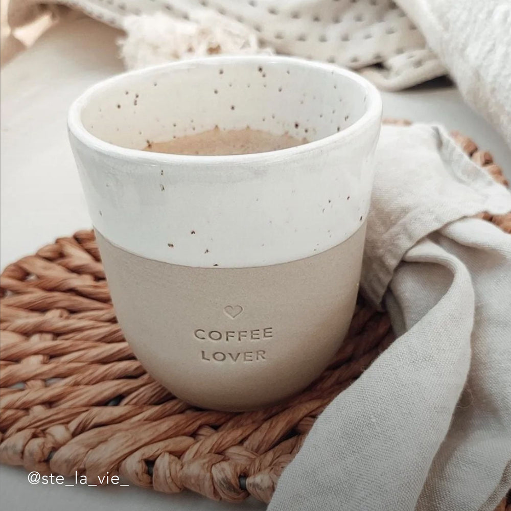 Ein Becher mit der Prägung "Coffee Lover" steht auf einem Ratan Untersetzer mit einer beigen Leinenserviette.