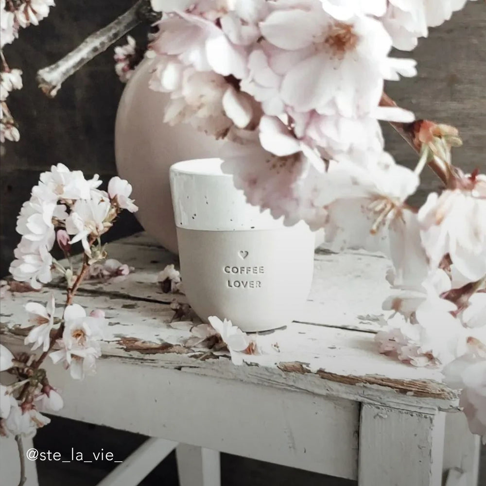 Ein Becher mit der Prägung "Coffee Lover" steht auf einem weißen shabby chic Holztisch mit einer Vase aus der wunderschöne rosa Blumen herausragen und alles umranken.