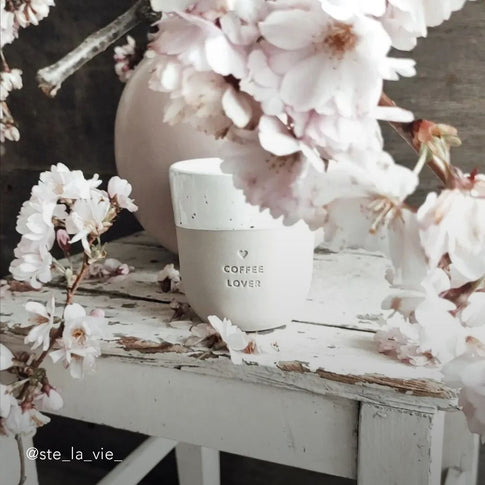Ein Becher mit der Prägung "Coffee Lover" steht auf einem weißen shabby chic Holztisch mit einer Vase aus der wunderschöne rosa Blumen herausragen und alles umranken.