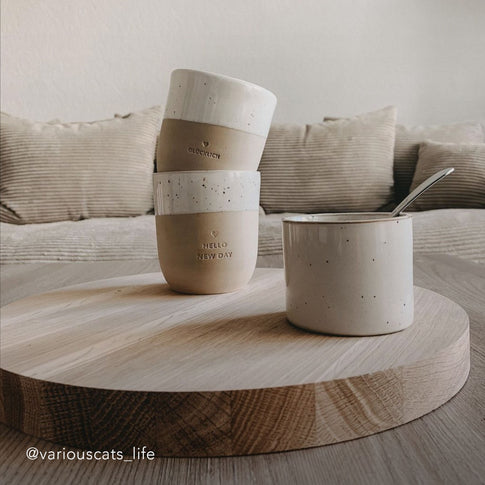 Zwei Keramikbecher mit Prägungen „GLÜCKLICH“ und „HELLO NEW DAY“ sind gestapelt, daneben ein weiterer heller Becher mit Löffel. Alle stehen auf einer runden Holzplatte vor einem gemütlichen Sofa. Unten links der Instagram-Name: @variouscats_life.