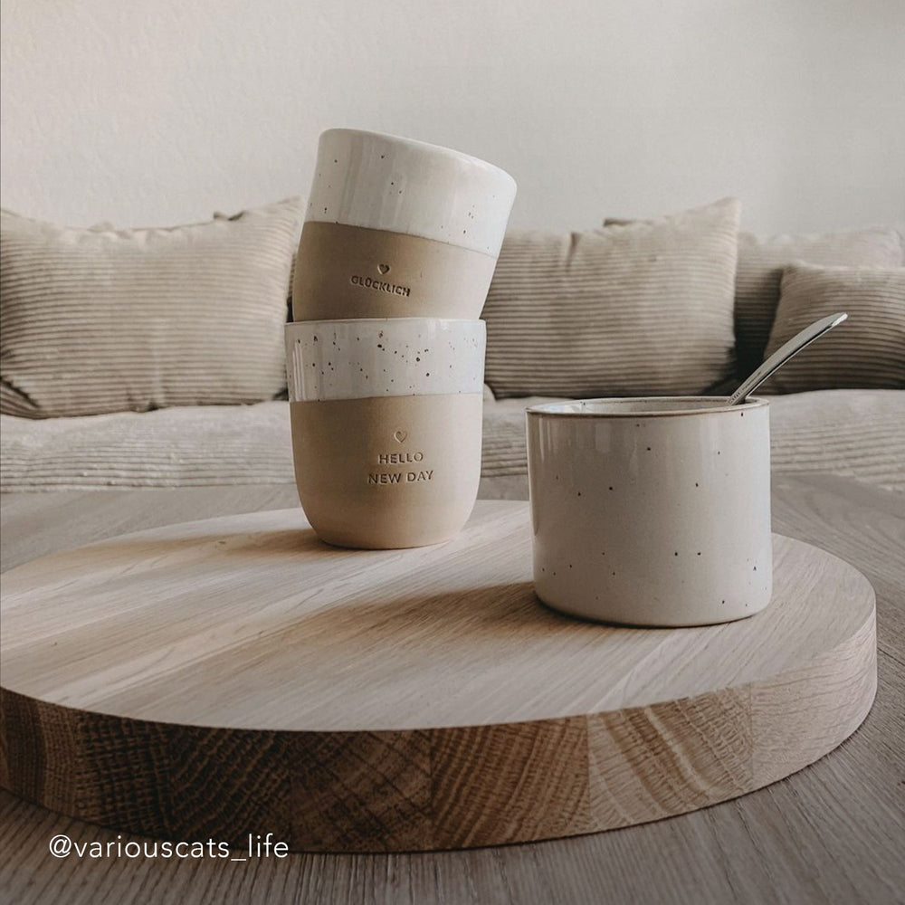 Zwei Becher und ein kleines Schälchen stehen auf einem Holztablett vor einem Sofa.