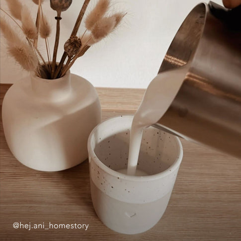 Becher Herz steht auf Holztisch, Milch wird eingeschänkt.