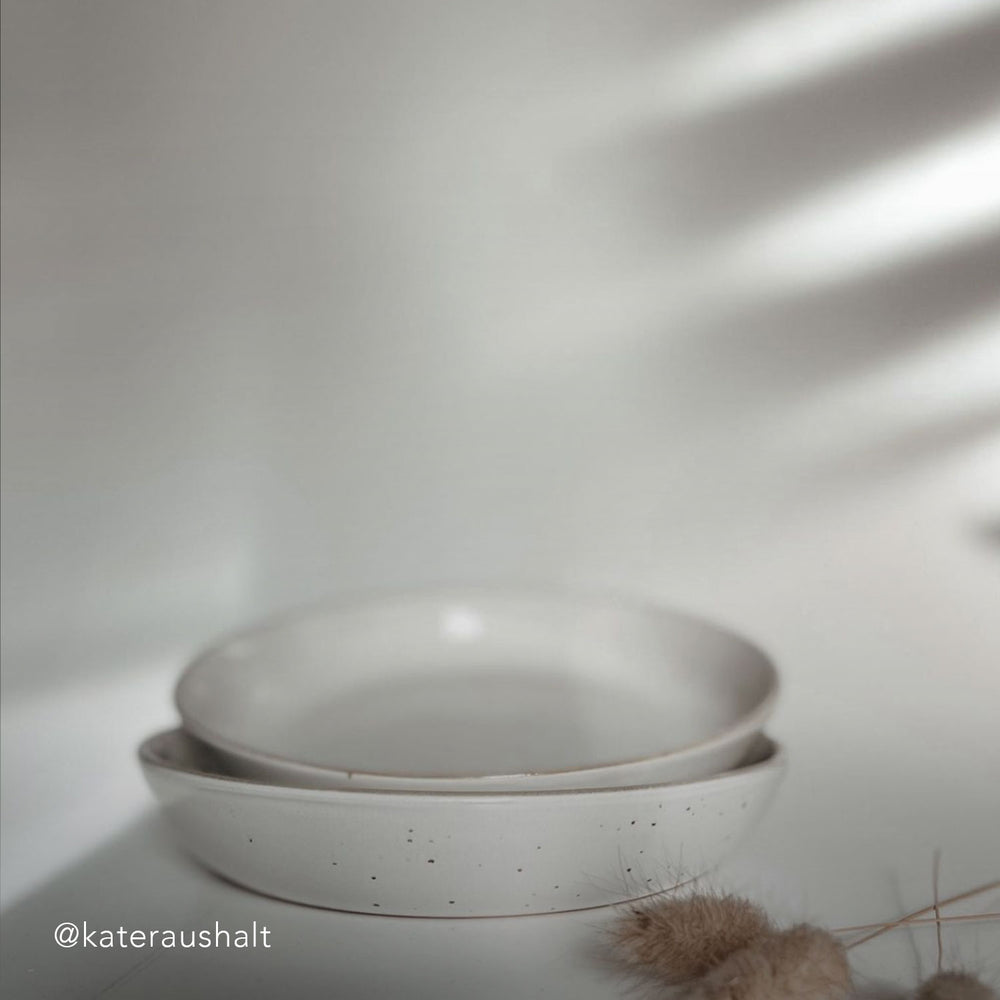 Zwei gestapelte, tiefe Teller in natürlicher Keramikoptik mit Sprenkelung liegen in weichem Licht auf einer hellen Fläche. Im Vordergrund getrocknete Gräser.