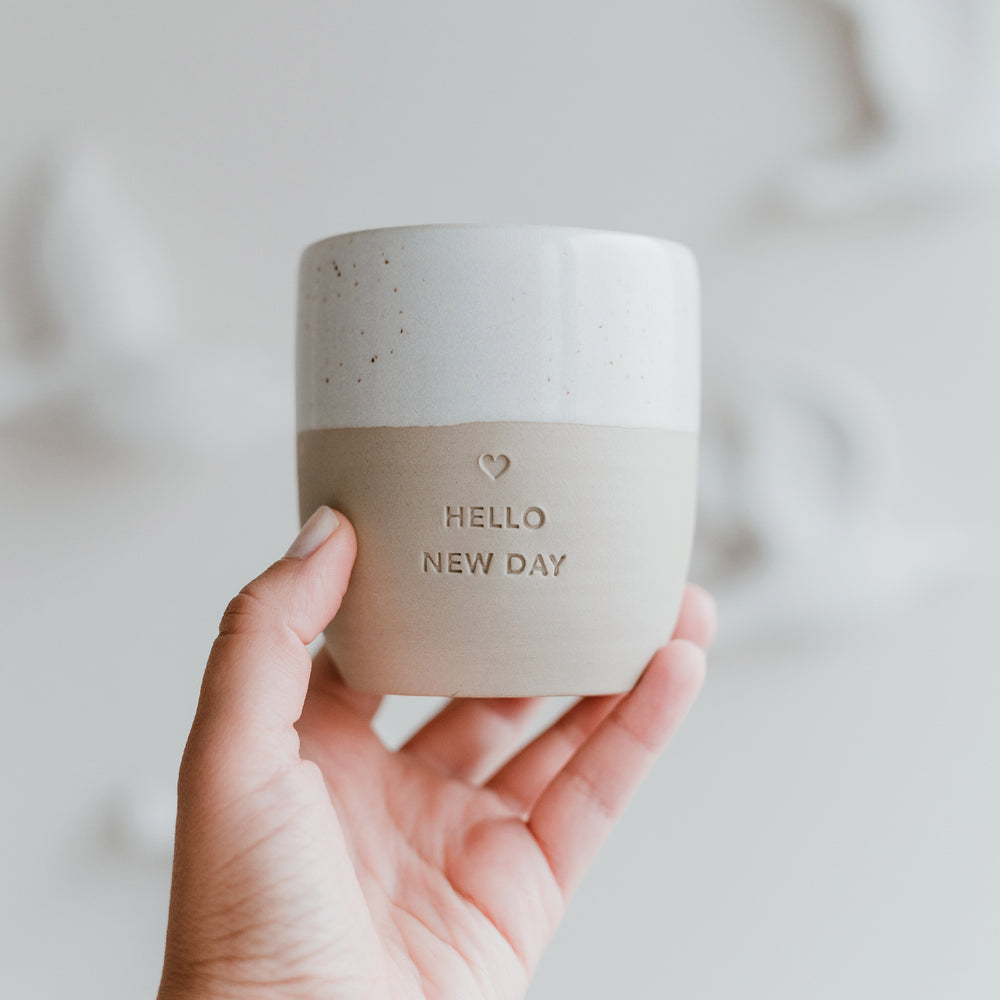 Eine Hand hält einen Becher mit der Prägung "Hello new day" mit einem mittigen Herz darüber.