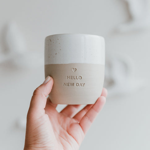 Eine Hand hält einen Becher mit der Prägung "Hello new day" mit einem mittigen Herz darüber.