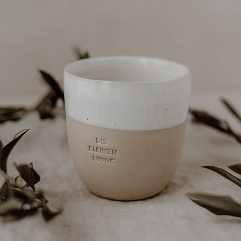 Ein einzelner Becher mit der Prägung ‚DIE KLEINEN DINGE‘ steht auf einem Leinentuch, sanft umrahmt von Olivenzweigen.